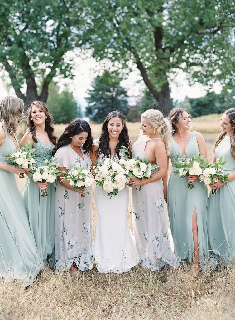 Bride and Bridesmaids in floral dresses in pastel green and lavender laugh while in a field in Discovery Park Seattle - Jacqueline Benét Photography