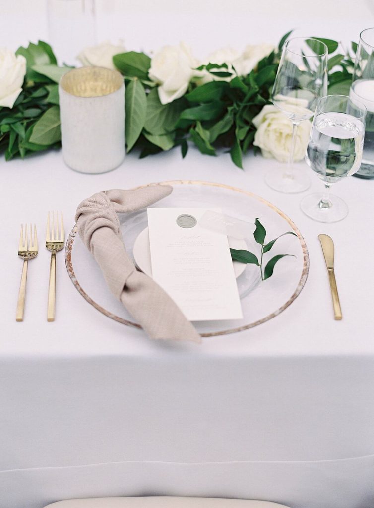 Gold and clear glass plate with white garland on wedding reception tabletop - Jacqueline Benét Photography