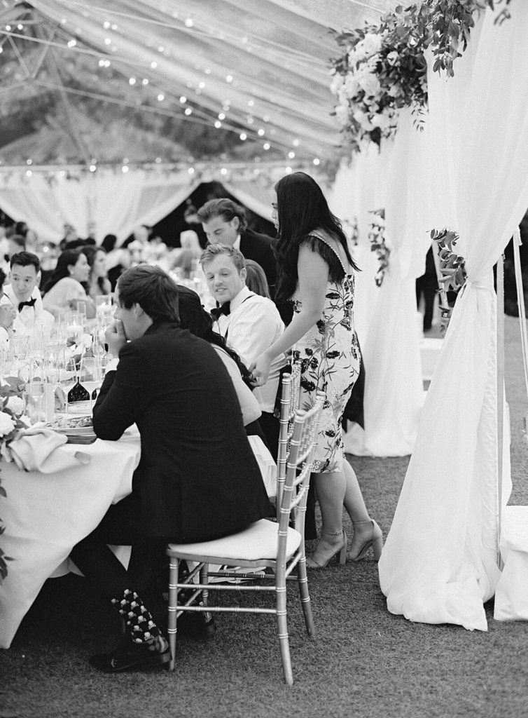 black and white photo of guests mingling under clear wedding reception tent - Jacqueline Benét Photography