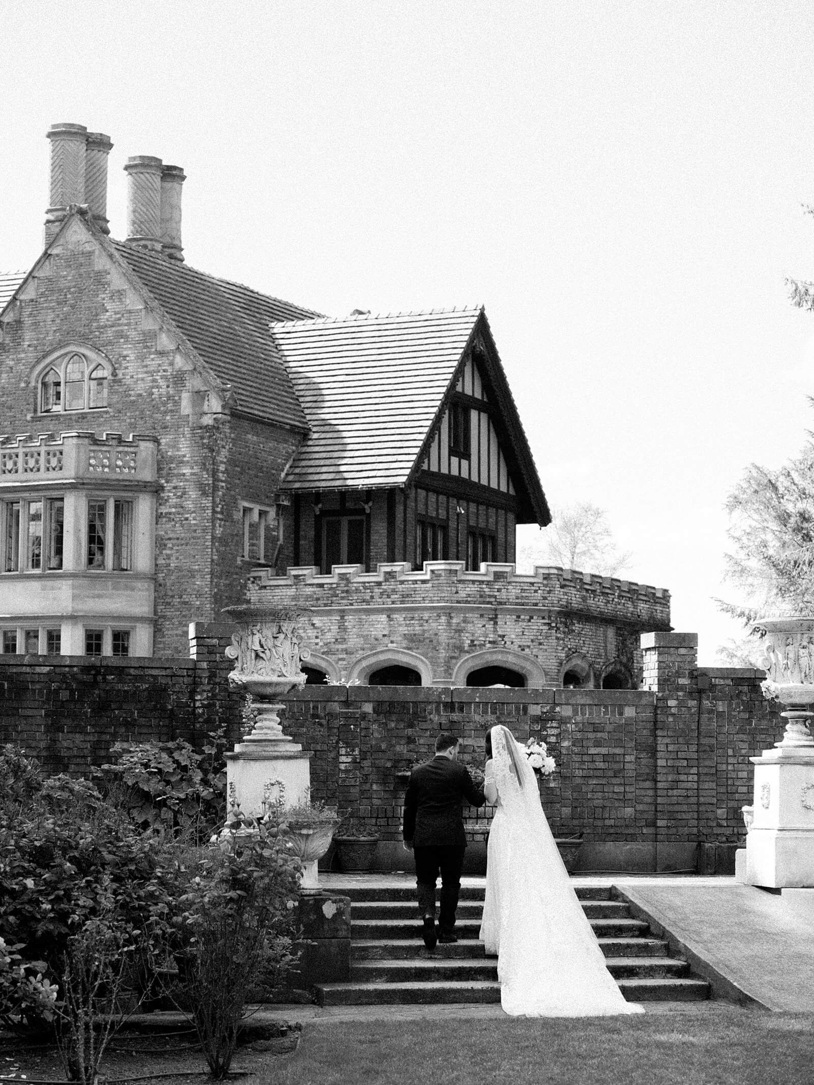 Bride and groom in black and white walk away up the steps toward Thornewood Castle - Jacqueline Benét Photography