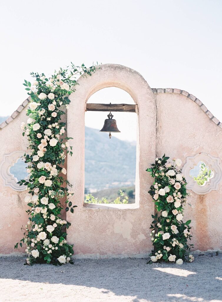 Blush floral arch flanking an old world bell on a pink stucco wall at Chateau Carmel - Jacqueline Benet Photography