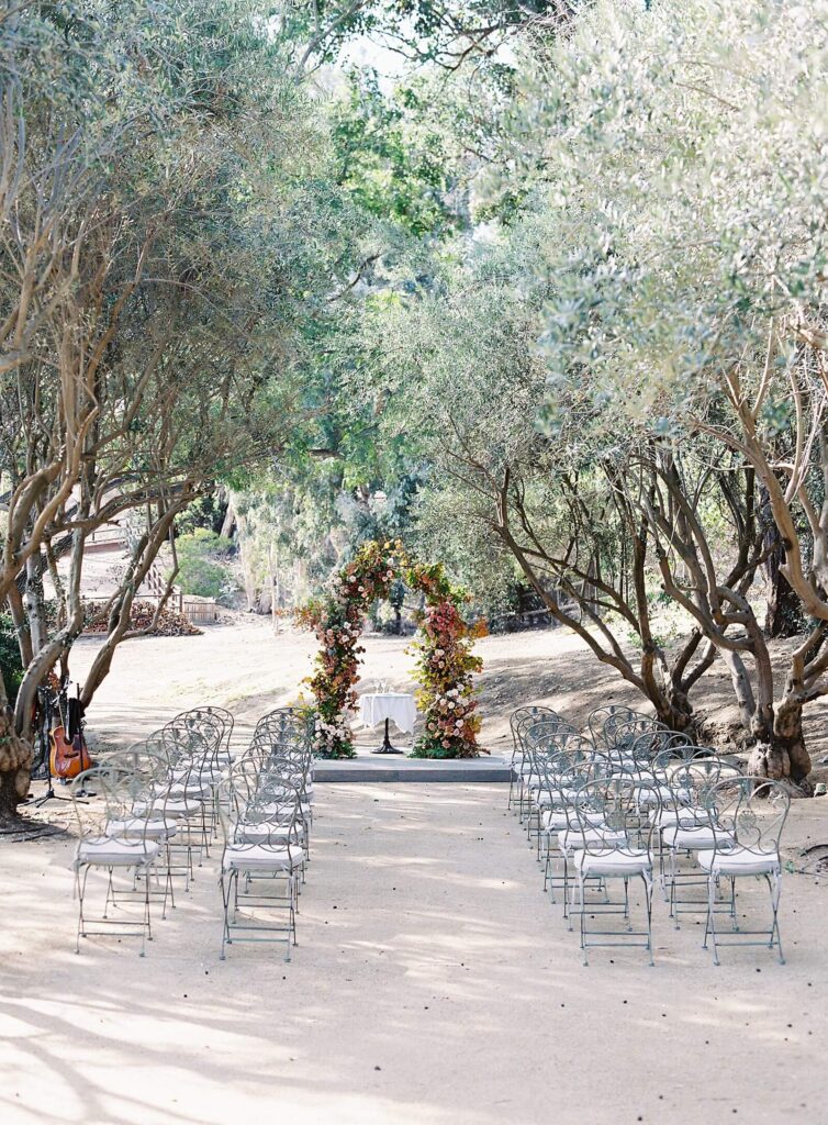 Fall pink red orange ombre floral arch with garden metal chairs at a wedding ceremony among olive grove - Jacqueline Benet Photography