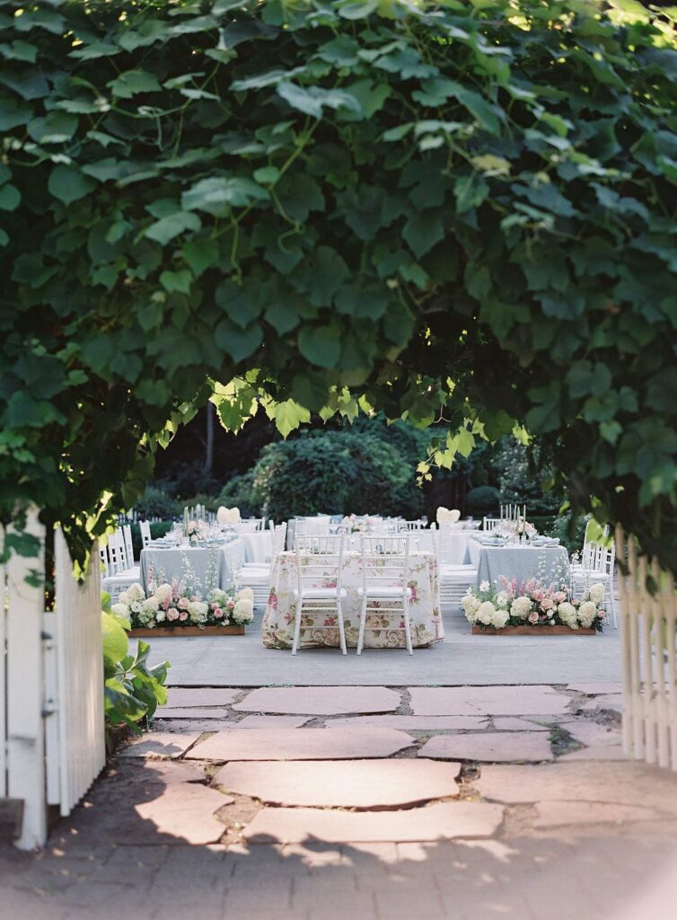 Wedding reception in pale blue and blush with white chivari chairs and floral tablecloth. Garden gates and arch in the foreground - Jacqueline Benét Photography