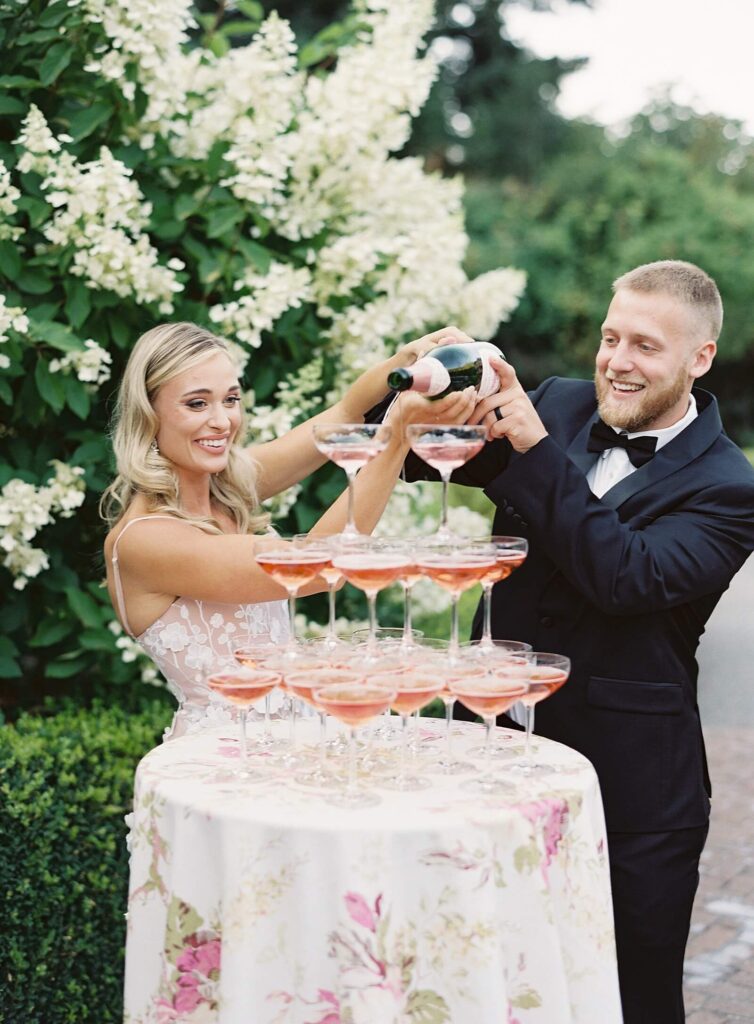 Bride and groom pour rose champagne at a champagne tower on floral tablecloth in front of hydrangeas  - Jacqueline Benét Photography