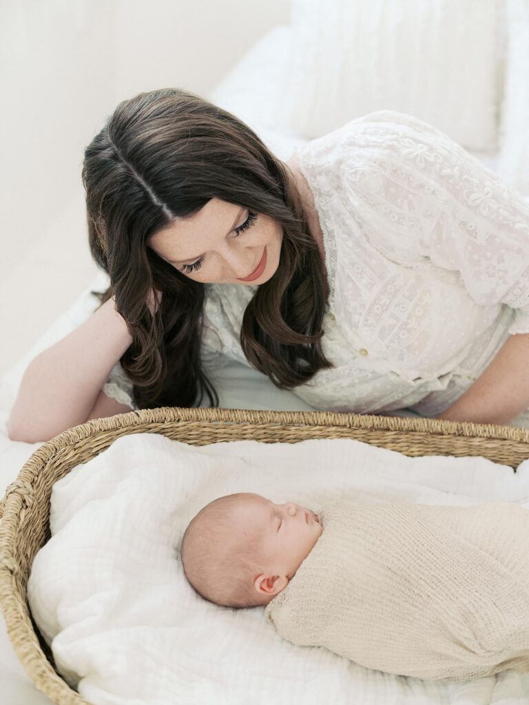 mom smiles down at newborn baby swaddled sleeping in a moses basket in photo taken in seattle studio