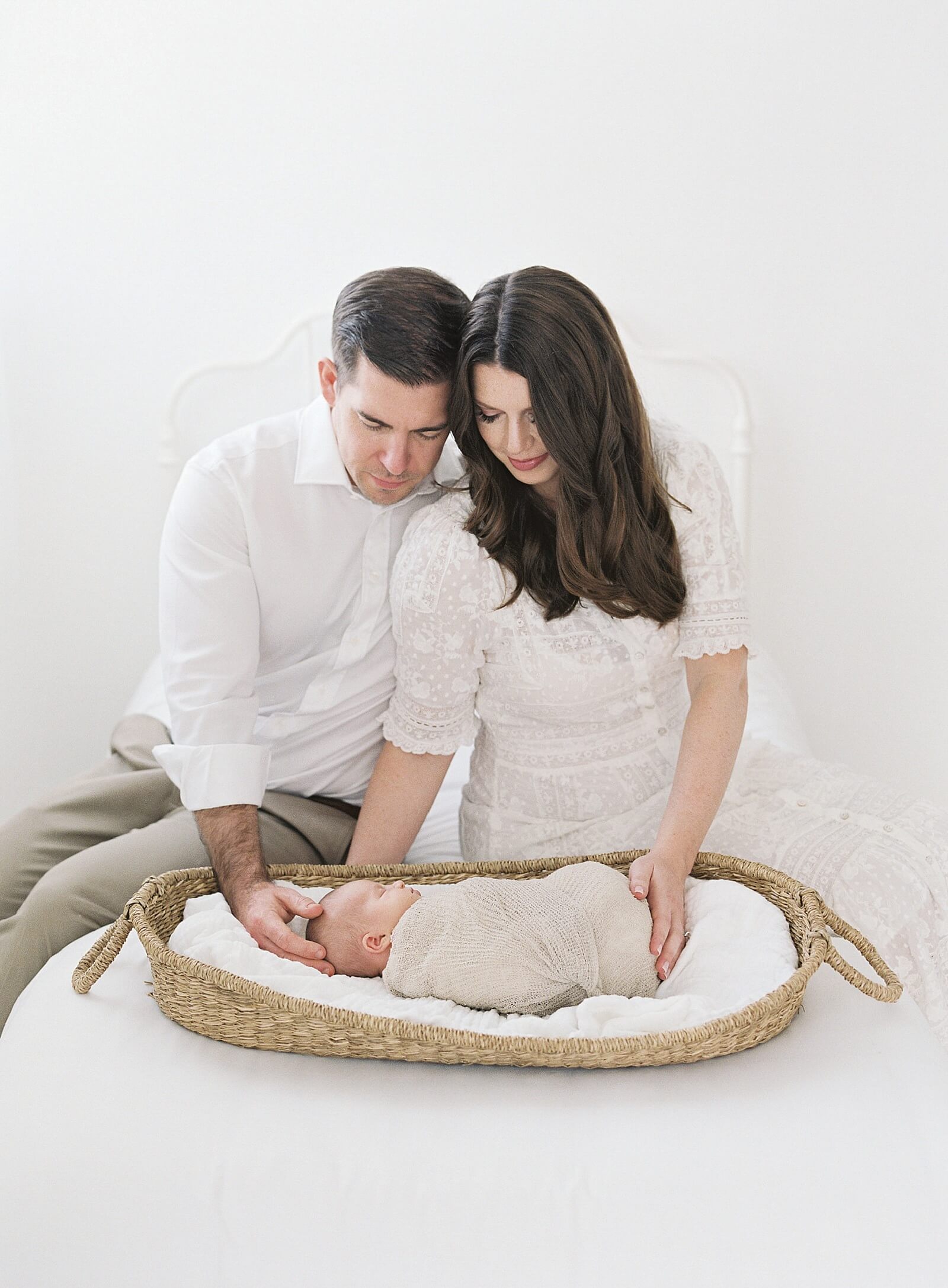 mom and dad smile down on newborn baby swaddled sleeping in a basket in an all white studio in Seattle taken by Jacqueline Benét