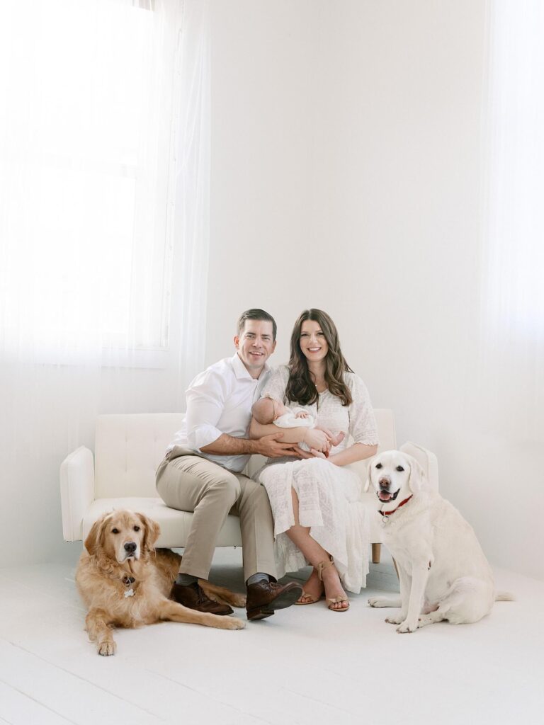 Mom and dad with newborn baby and two dogs smile at the camera in all white Seattle studio with photographer Jacqueline Benét