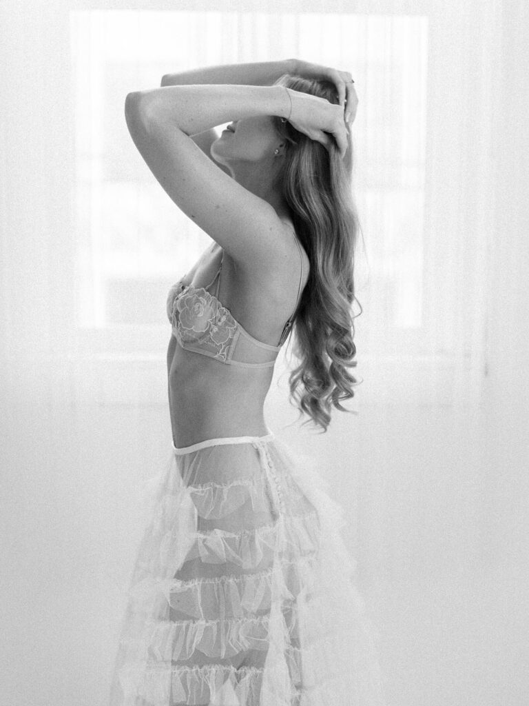 Black and white photo of a woman standing in profile with hands in her hair, wearing lace lingerie and a sheer skirt during a birthday boudoir photoshoot in Seattle.