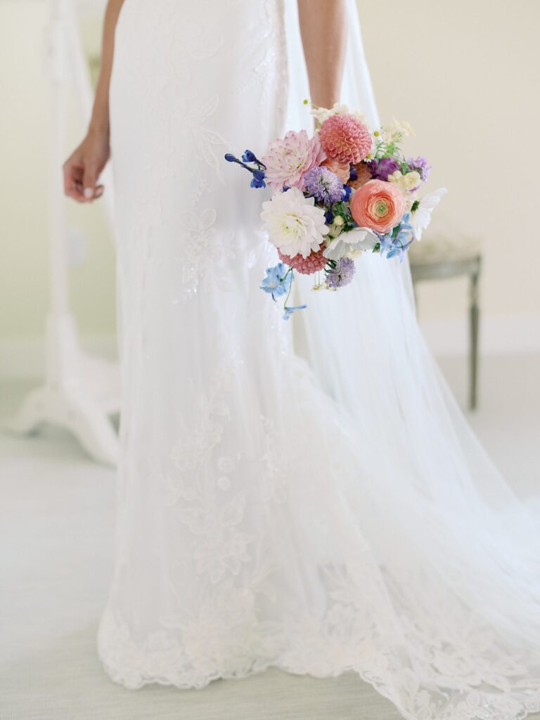 Detail of bride’s lace wedding gown and colorful bouquet featuring ranunculus, dahlias, and delphinium at a Harmony Meadows wedding.