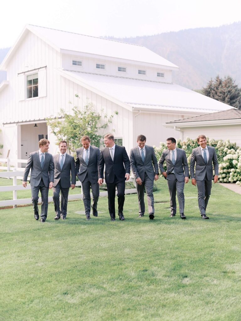Groom walking with his groomsmen in gray suits outside the white barn at Harmony Meadows wedding venue.