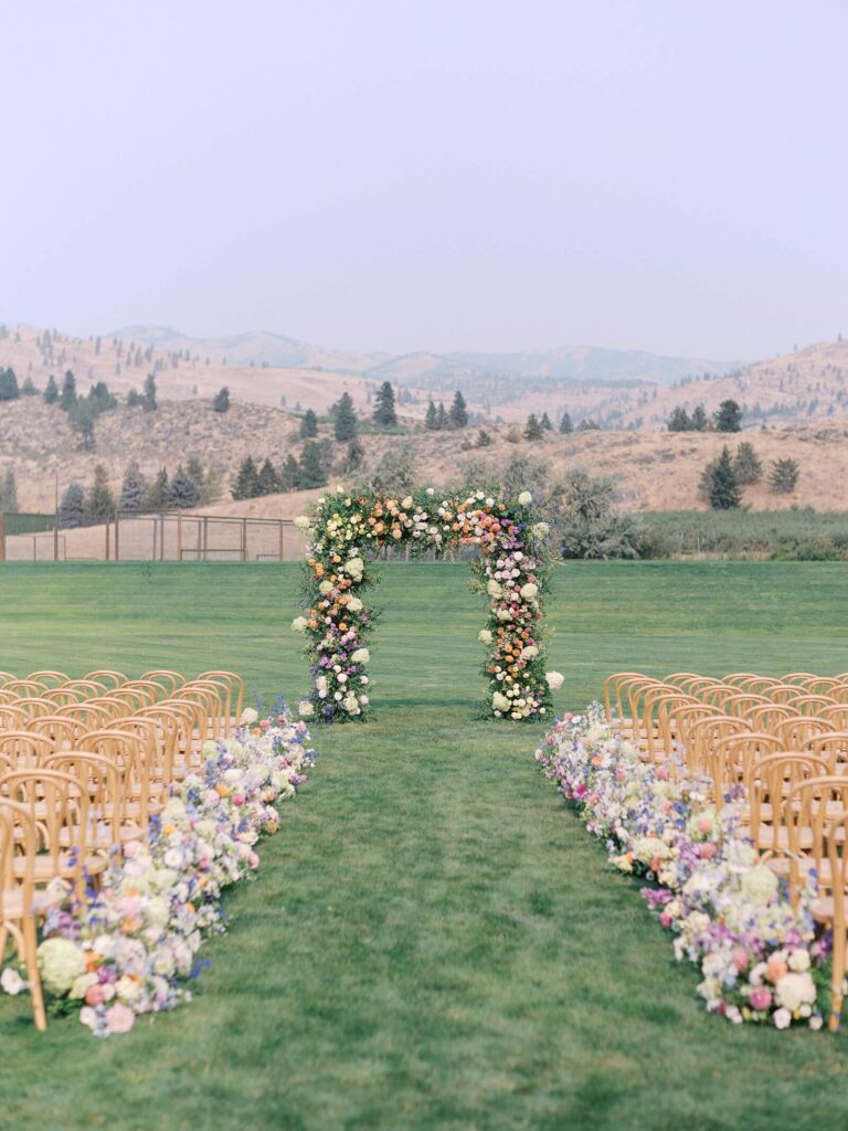 Ceremony setup with wooden chairs, floral aisle arrangements, and a colorful flower arch overlooking the mountains at a Harmony Meadows wedding.