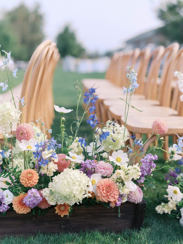 Close-up of colorful floral arrangements lining the ceremony aisle with wooden chairs set up for an outdoor Harmony Meadows wedding.