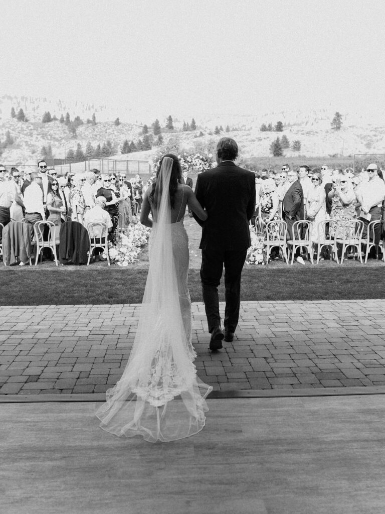 Black and white photo of the bride walking down the aisle arm-in-arm with her father as guests look on during a Harmony Meadows wedding ceremony.