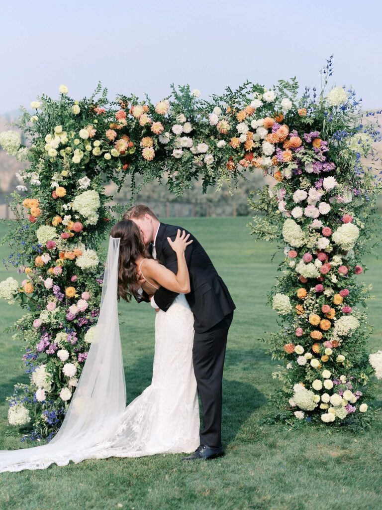 Bride and groom sharing a kiss beneath a lush, colorful floral arch during their outdoor Harmony Meadows wedding ceremony.