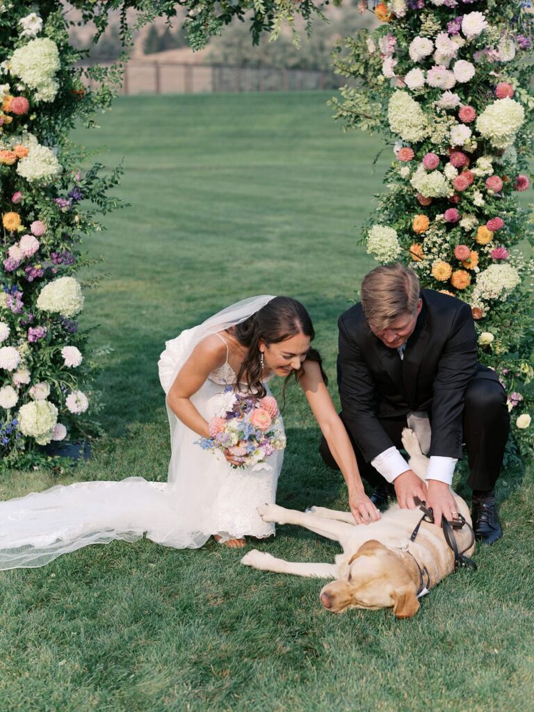 Bride and groom smiling and petting their dog beneath a lush floral ceremony arch during their Harmony Meadows wedding.