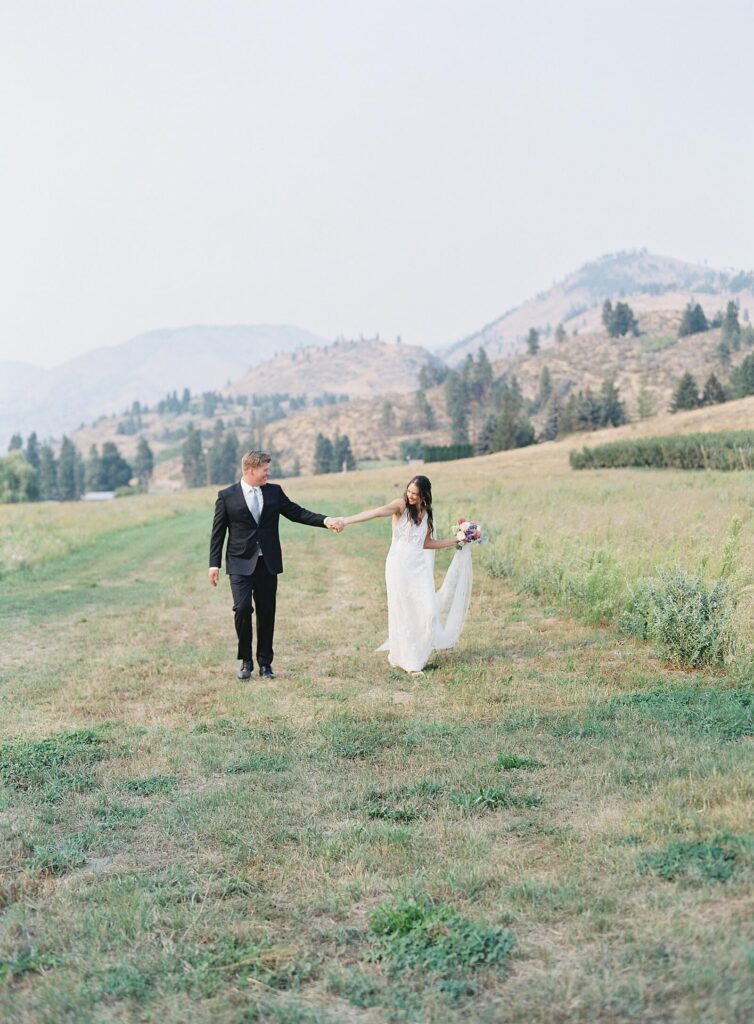 Bride and groom walking hand in hand through a sunlit mountain field at their Harmony Meadows wedding.