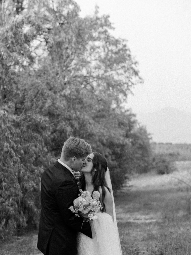 Black and white photo of bride and groom sharing a romantic kiss in a wooded field at their Harmony Meadows wedding.
