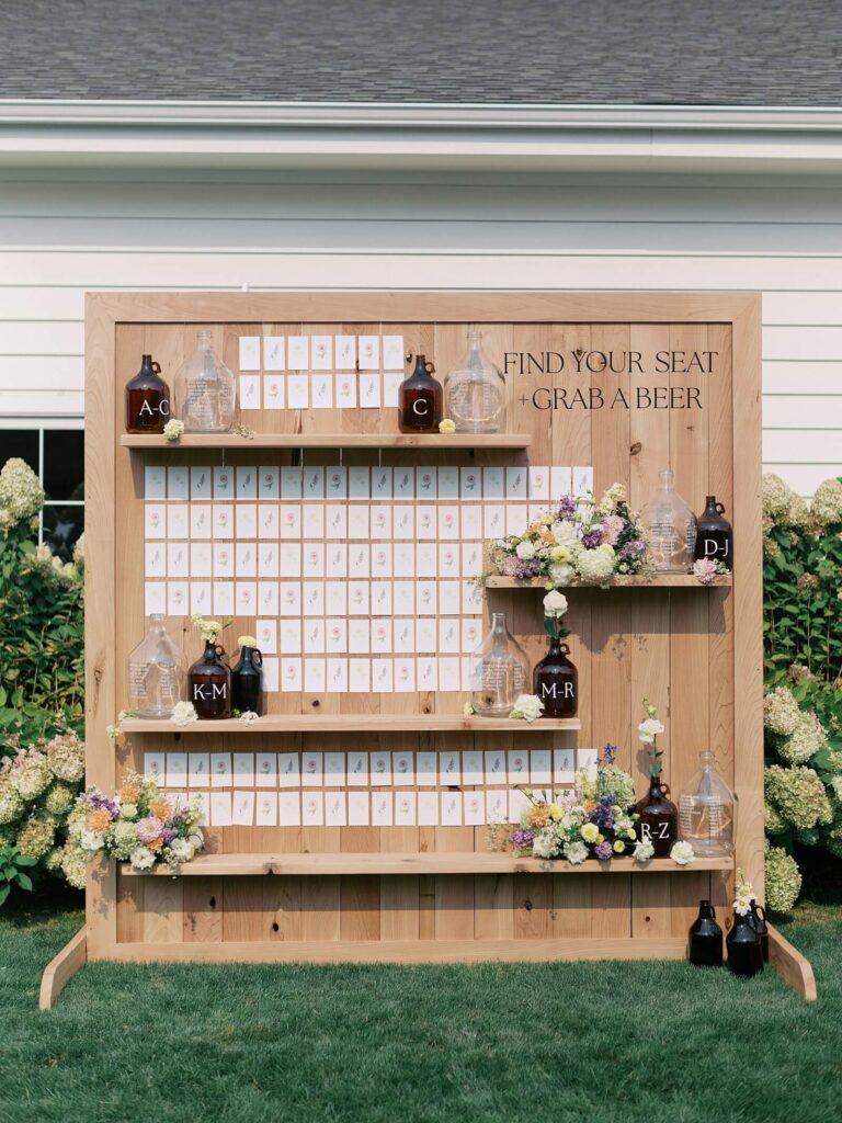 Creative wooden escort card display with floral arrangements and beer growlers at a Harmony Meadows wedding.