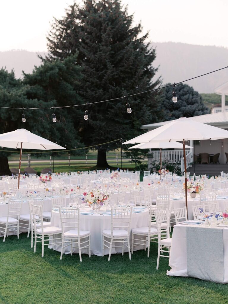 Outdoor reception setup with round tables, white chairs, umbrellas, and colorful floral centerpieces at a Harmony Meadows wedding.