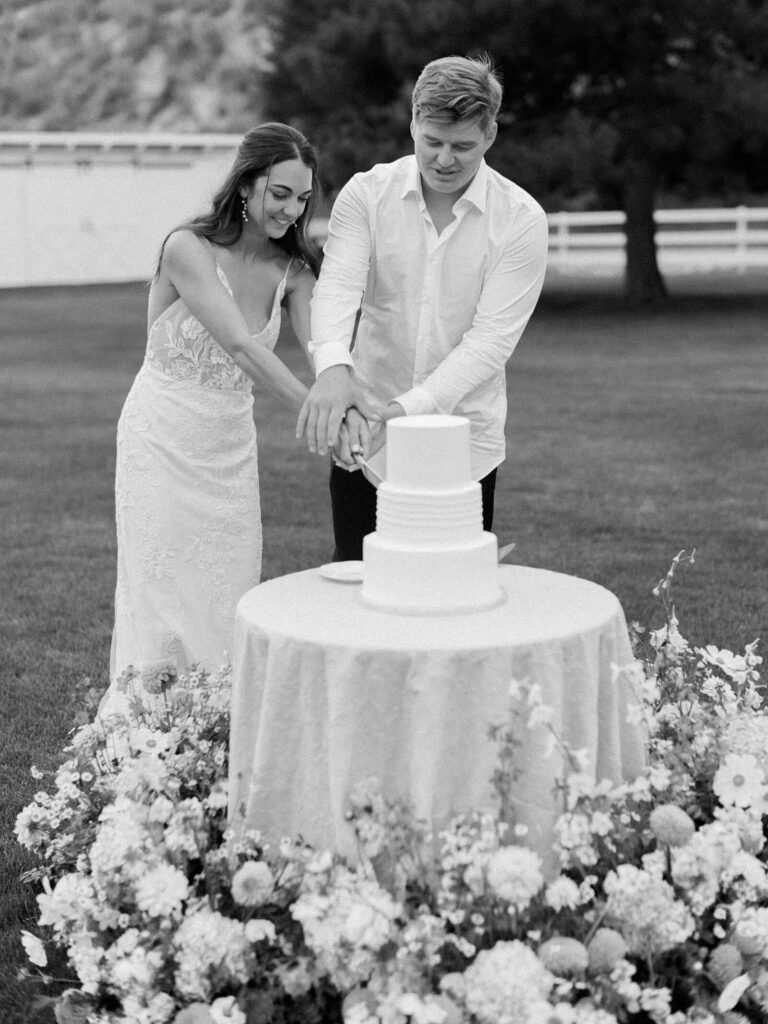 Black and white photo of bride and groom cutting their wedding cake surrounded by flowers during their Harmony Meadows wedding reception.