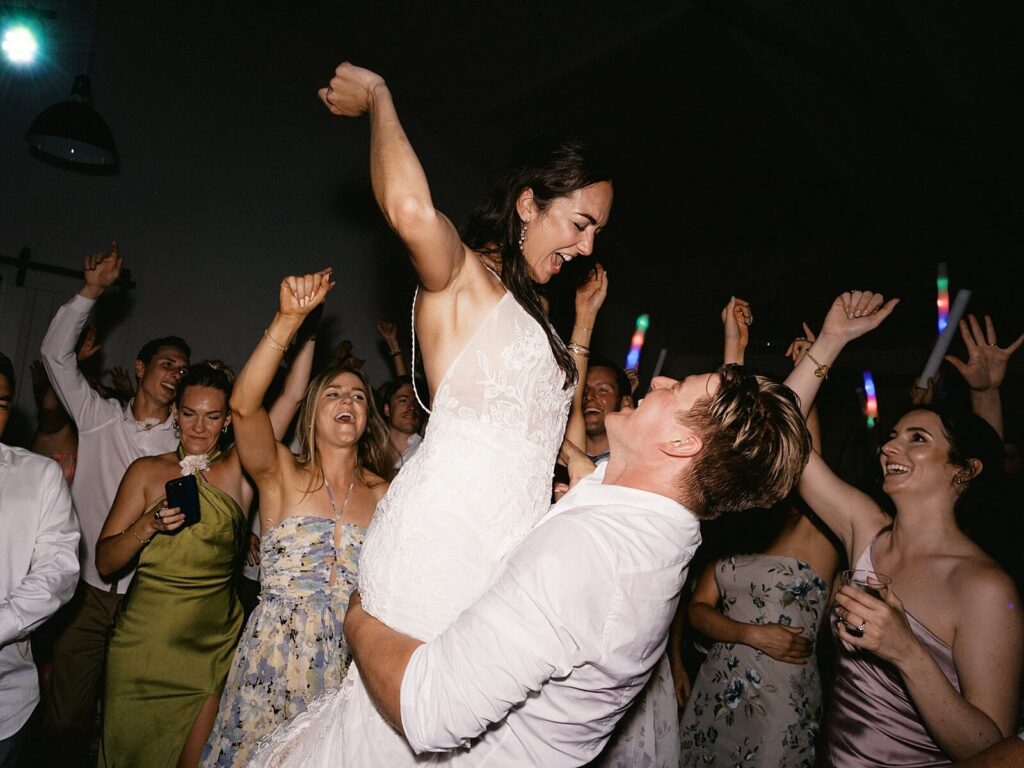 Bride and groom celebrating on the dance floor surrounded by cheering guests with glow sticks during their Harmony Meadows wedding reception.