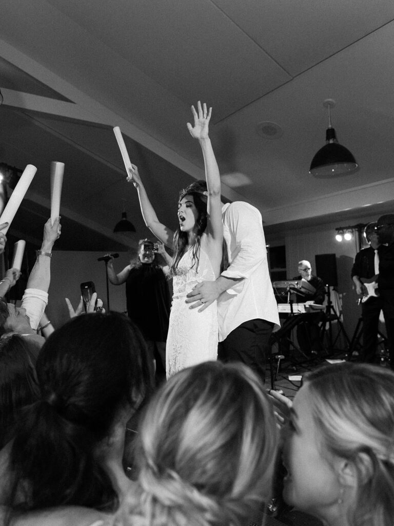 Black and white photo of the bride cheering with glow sticks on the dance floor during a lively Harmony Meadows wedding reception.