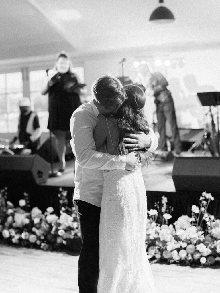 Black and white photo of bride and groom embracing during their first dance as a live band performs at their Harmony Meadows wedding reception.