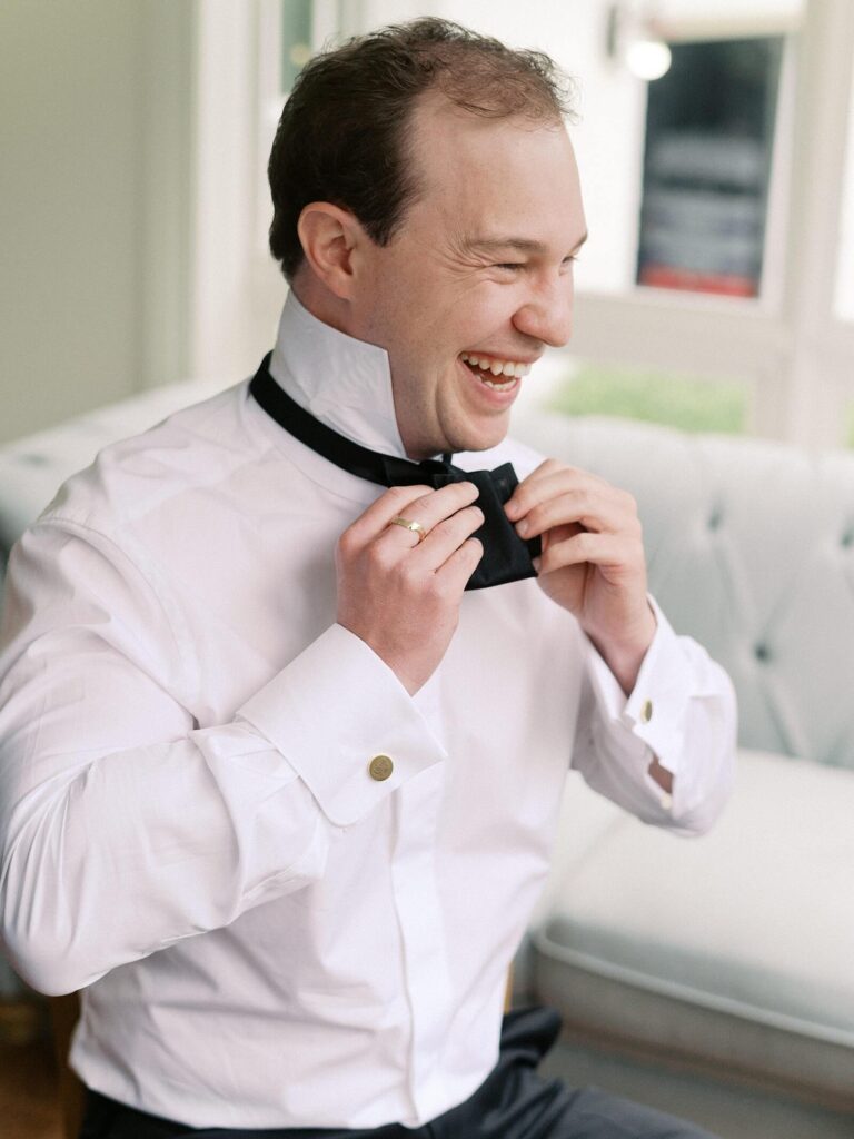 The groom putting smiling while putting on his bowtie as he prepares for his garden wedding at Fox Hollow Farm, one of Seattle best outdoor wedding venues - by Jacqueline Benét 
