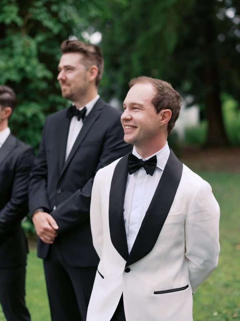 A close up of the groom smiling and watching his bride walk towards him down the ceremony isle. Captured by Jacqueline Benét | Fine art wedding photography 