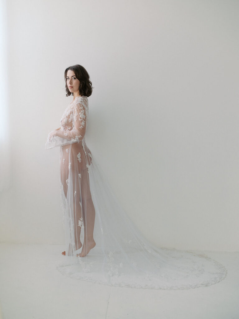 Bridal boudoir photo of a woman standing in bright light, wearing a long, lacy and sheer robe. She is looking at the camera with a smoldering look. Taken by a Seattle boudoir photographer. 